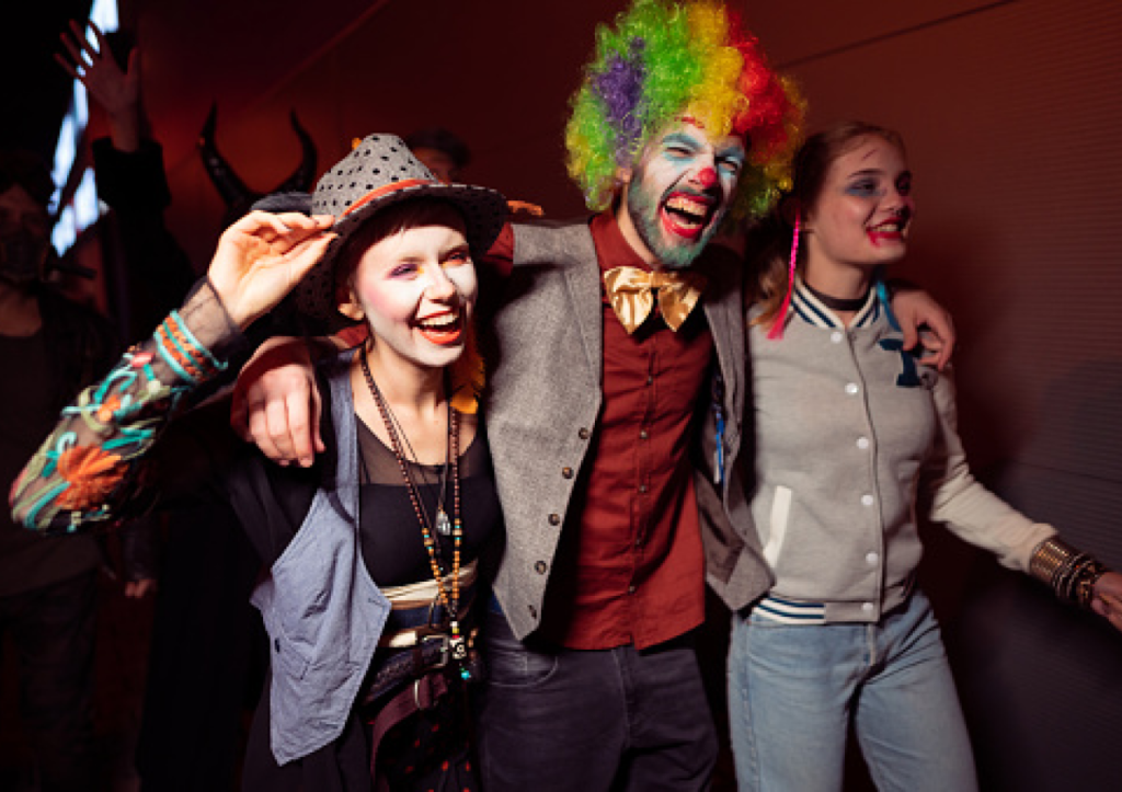 Three friends laughing in halloween costumes (hipster, clown, zombie teen) with their arms over each others shoulders